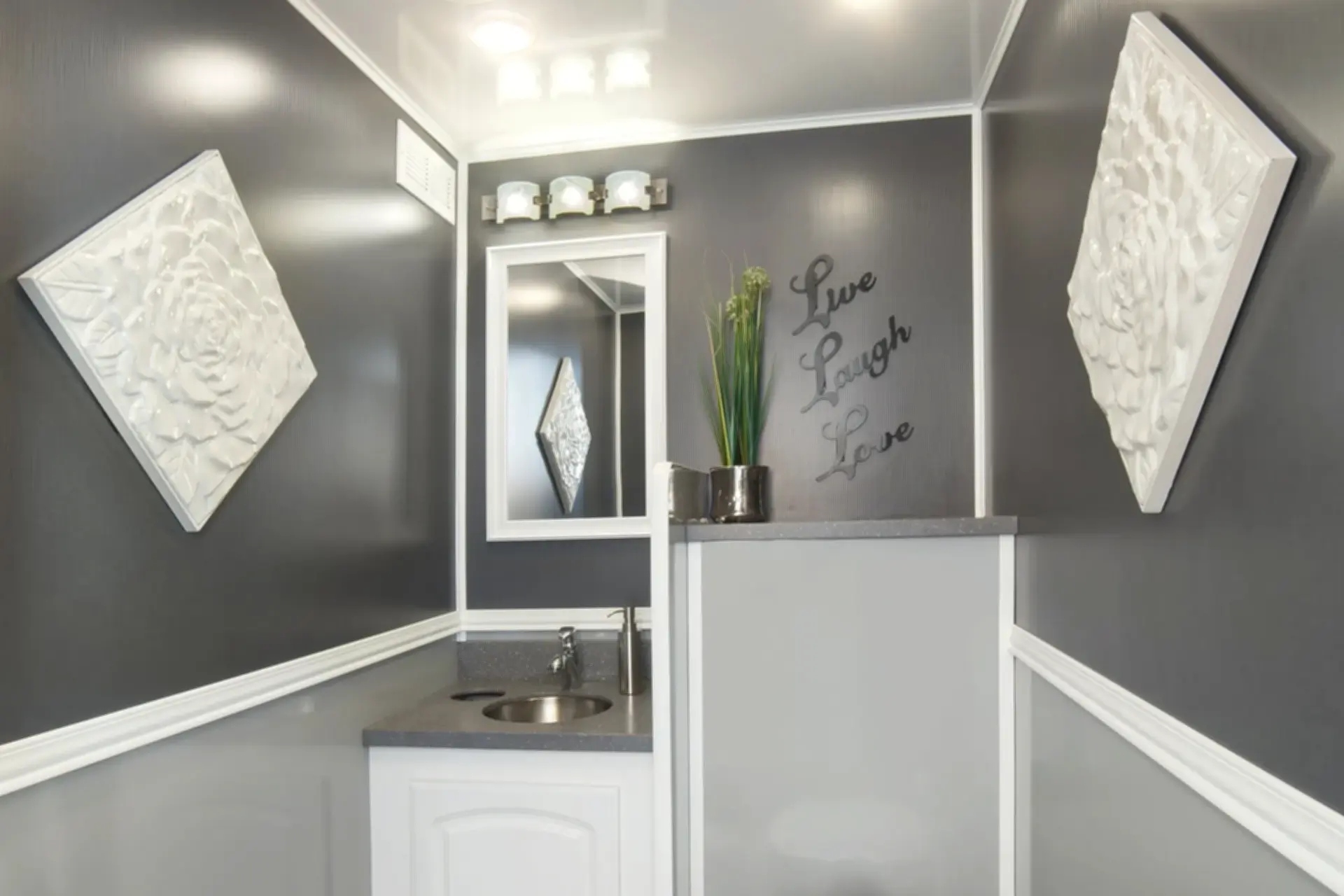 Restroom Trailer featuring modern gray walls, white decorative wall art, a sink with a faucet, and a mirror, complemented by a Live Laugh Love wall quote and a plant on a countertop.