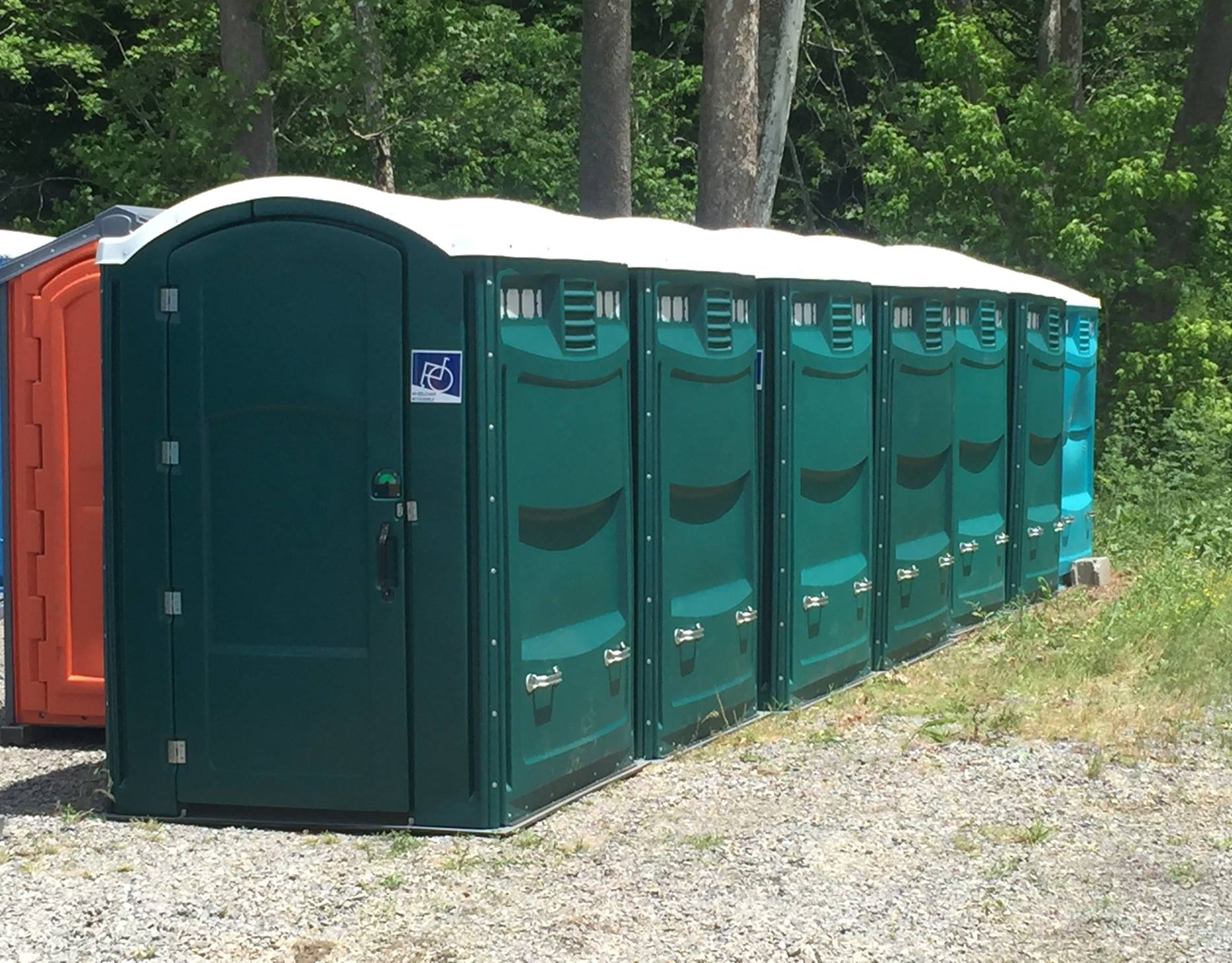 A row of green portable toilets situated on gravel, with trees and greenery in the background. One unit has a handicap accessibility sign.