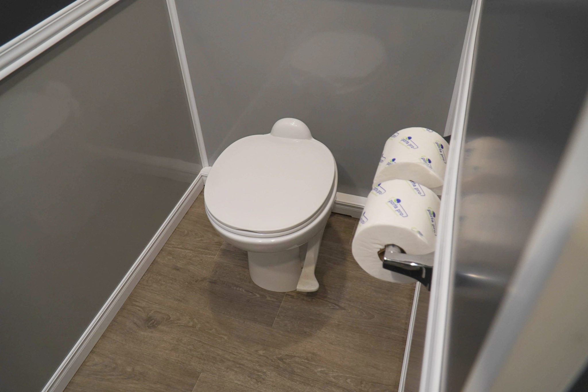 Portable Toilet interior featuring a white toilet and two rolls of toilet paper on a holder. The walls are light gray with a polished wooden floor.
