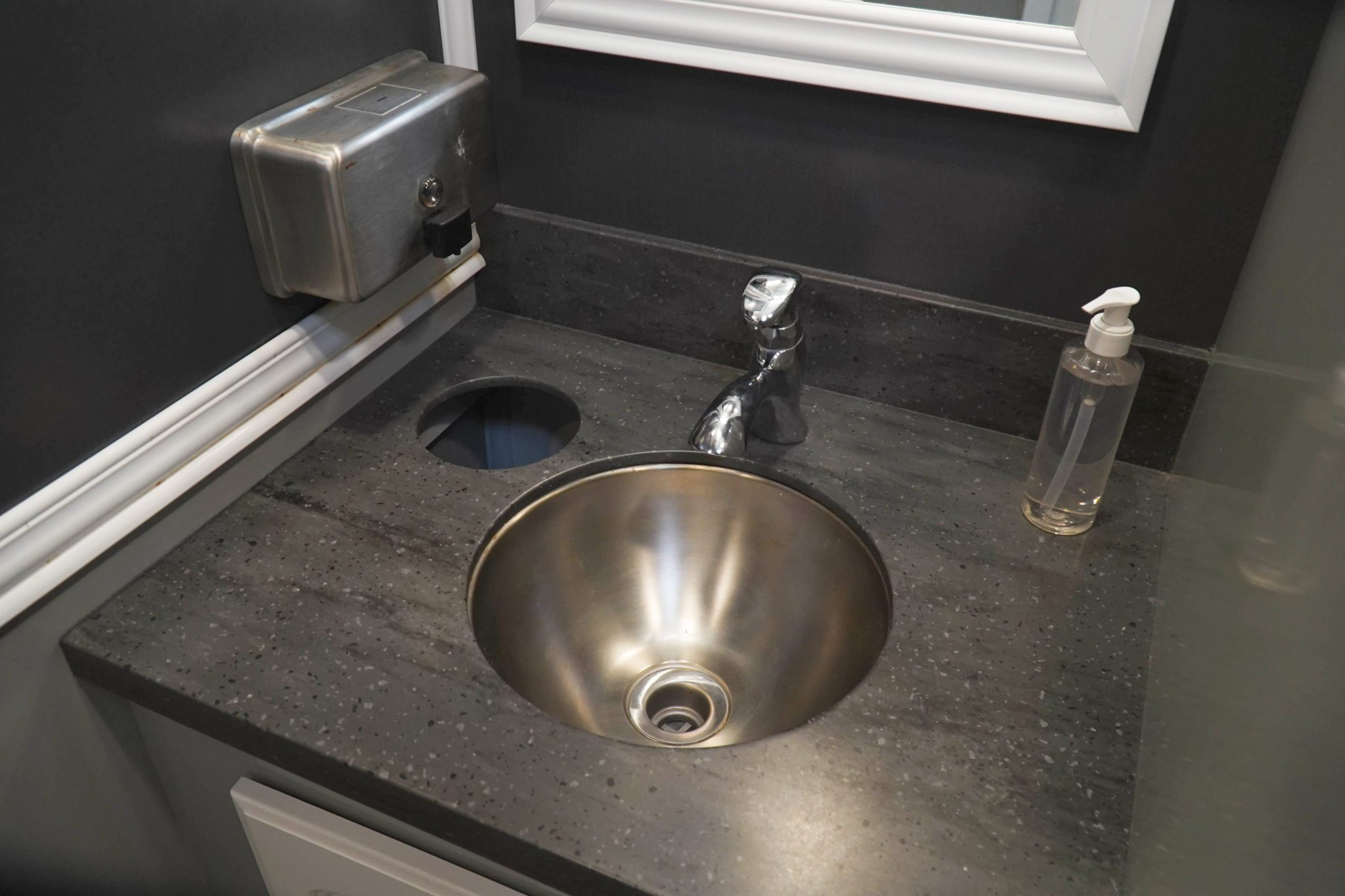 Interior view of a restroom with a stainless steel sink and faucet on a dark speckled countertop. A clear soap dispenser is placed next to the sink, and above it is a silver paper towel dispenser mounted on the wall. A round hole in the countertop suggests additional functionality.